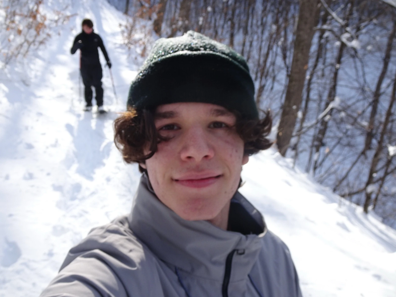 Ömer in a dark beanie on a snowed-in forest trail, a cross-country skier climbing behind him